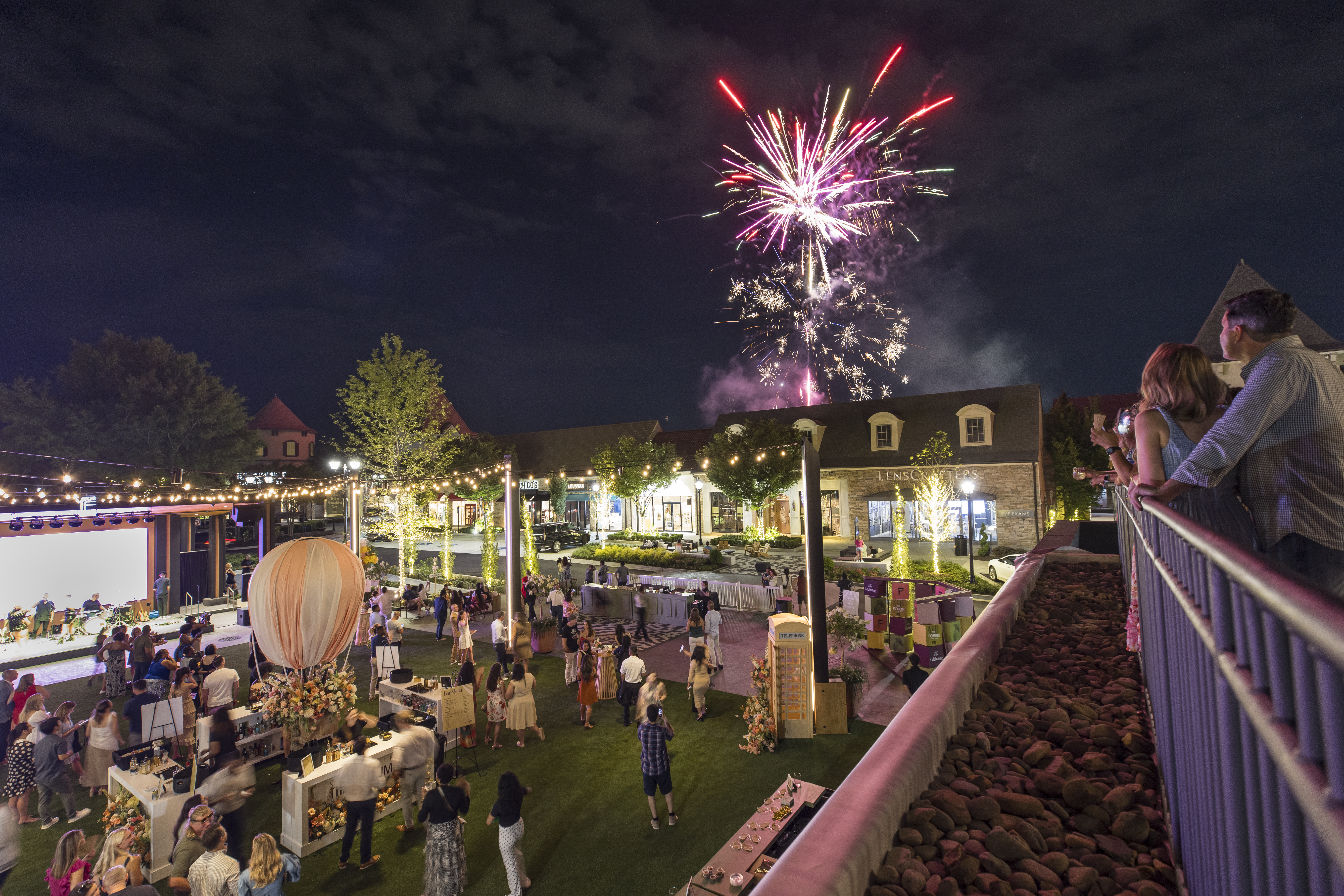 A group of people enjoying a lively outdoor event with colorful lights and live music, looking at fireworks in the night sky