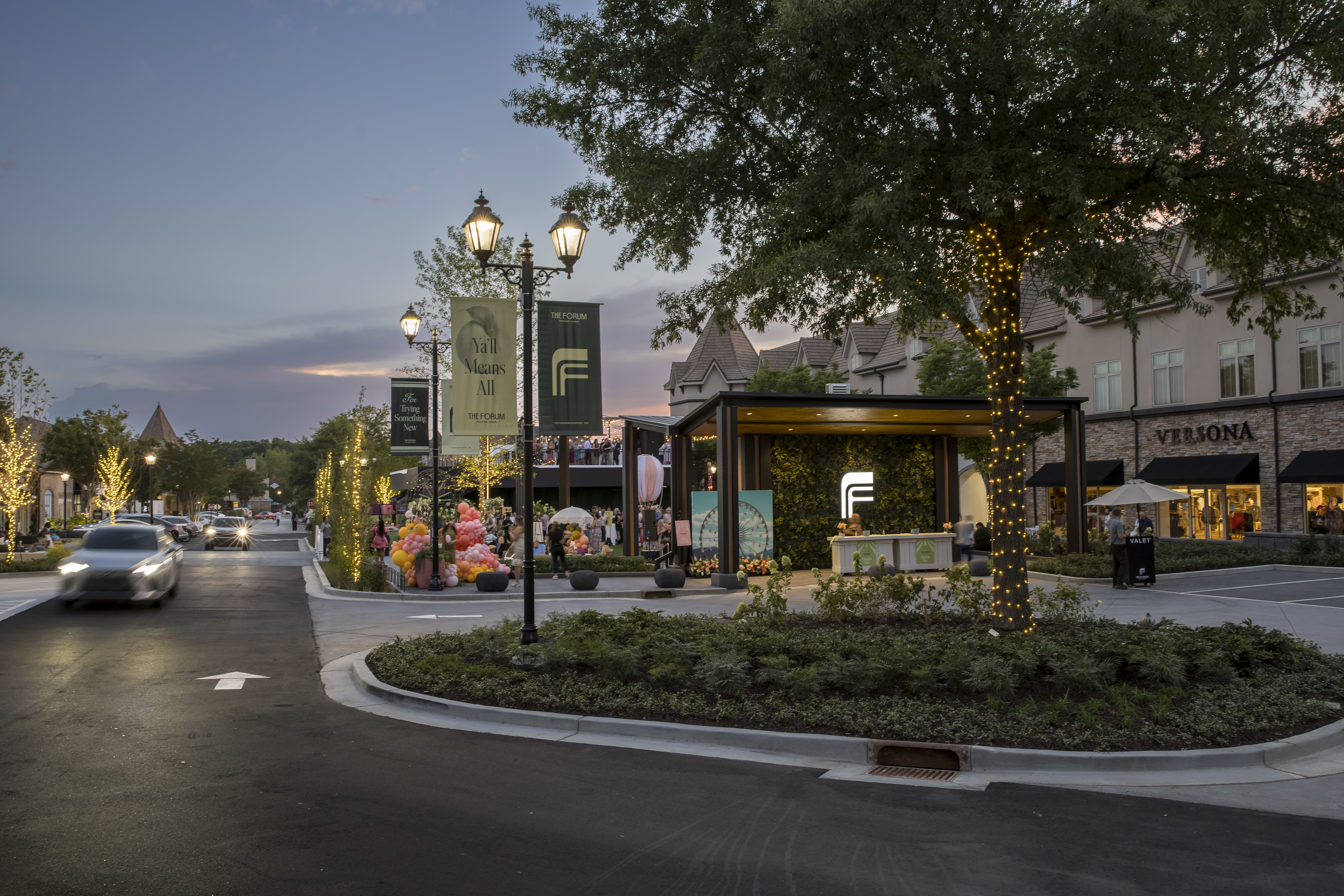 Evening view of a road at The Forum Plaza with festive tree lighting, cars driving, and a valet person in front of the Versona store.